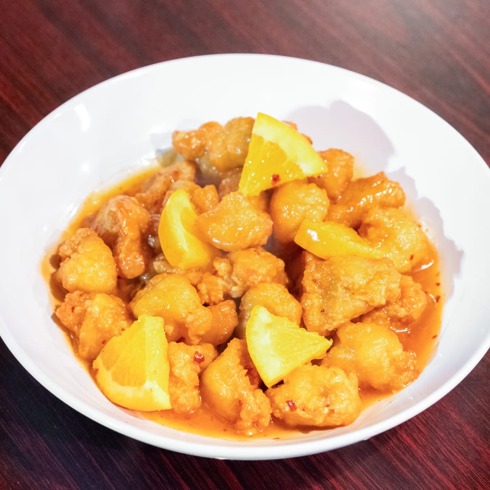Crispy golden chicken pieces in orange sauce garnished with fresh orange wedges, served in a white bowl on a wooden table.