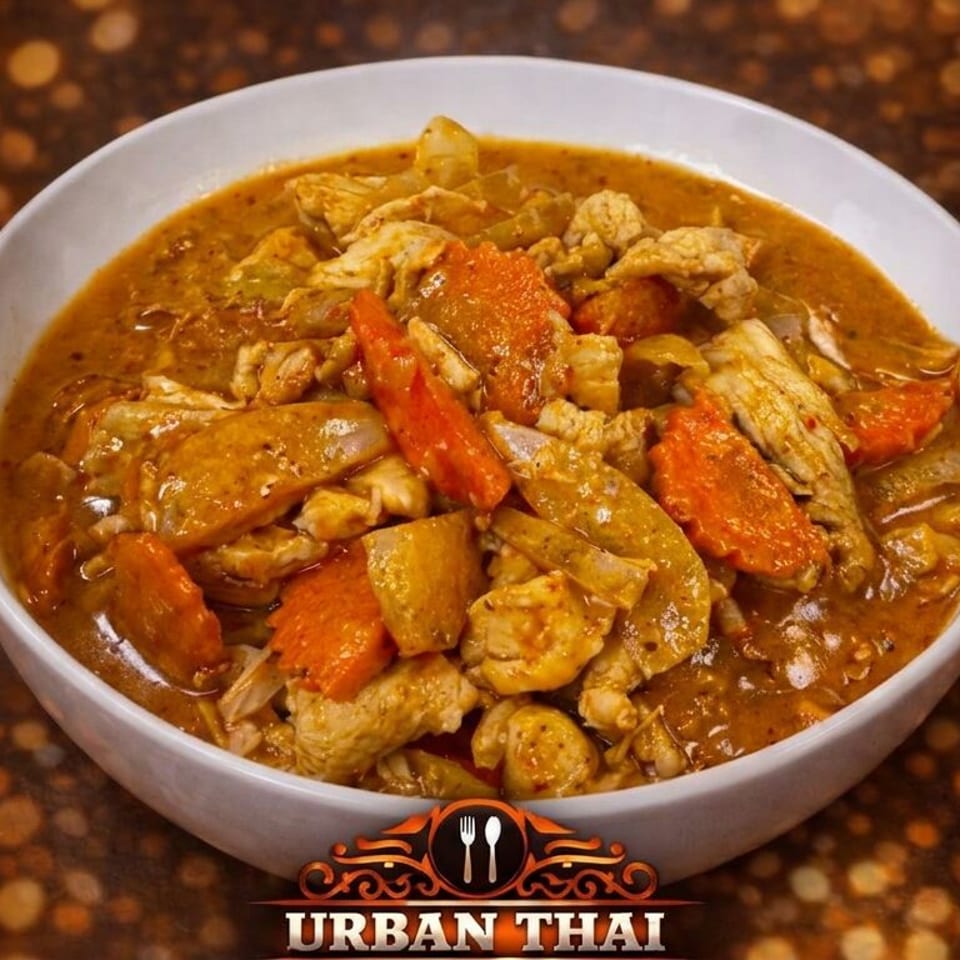 Bowl of Thai curry with chicken, carrots, onions, and vegetables on a dark speckled table, branded Urban Thai.