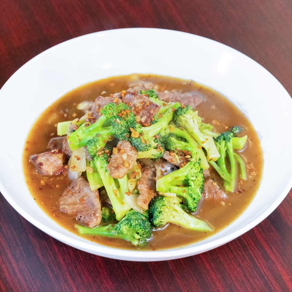 Beef and broccoli stir-fry in brown sauce, garnished with crispy garlic, served in a white bowl on a wooden table.