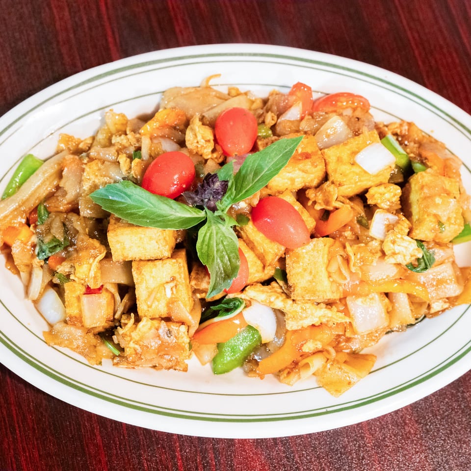 Thai stir-fried noodles with tofu, vegetables, scrambled egg, cherry tomatoes, and fresh basil on a plate.