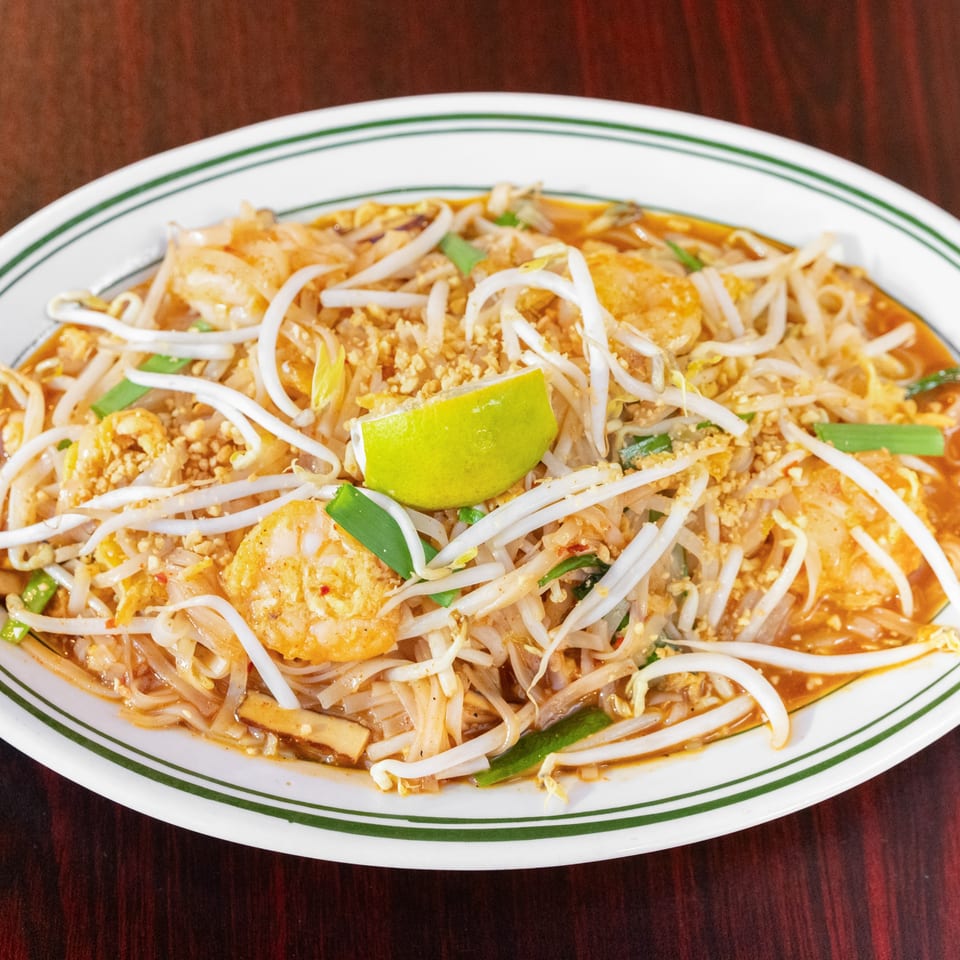Pad Thai noodles with shrimp, bean sprouts, crushed peanuts, scallions, and lime on a white platter.